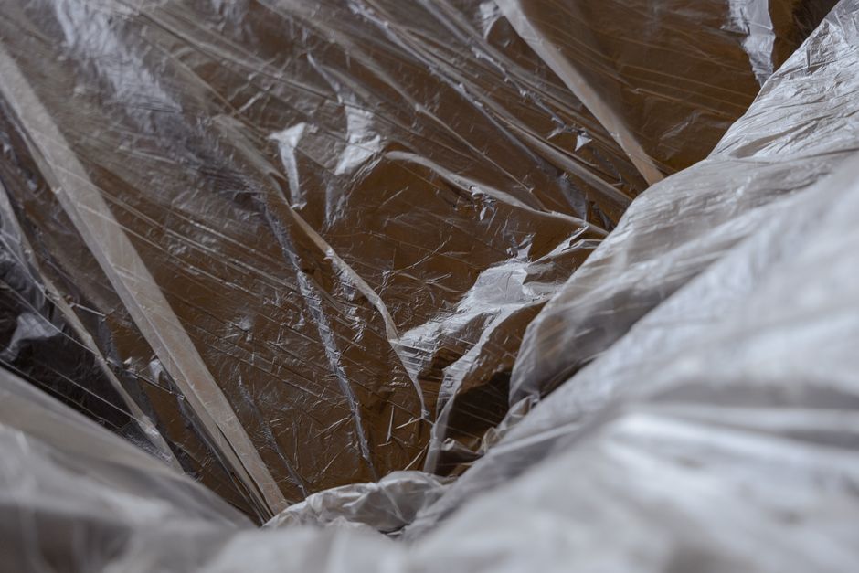 A close-up view of a large, transparent plastic rubbish bag filled with dark brown and black debris, possibly old wooden or cardboard materials, with a crinkled texture and reflective sheen, placed on a paved outdoor surface. In the background, additional plastic bags and scattered waste can be seen, indicating a small-scale rubbish removal activity near a property or clearance site. The environment suggests an area prepared for collection or disposal, typical of private rubbish clearance services operating independently from local authority waste management. Lighting appears natural, highlighting the varying textures and tones of the plastic and debris. This scene visually relates to the process of rubbish collection or on-site clearance, underscoring the importance of professional waste removal to manage excess loft or household disposal efficiently, aligning with services offered by Rubbish Clearance Kingston for general waste handling.