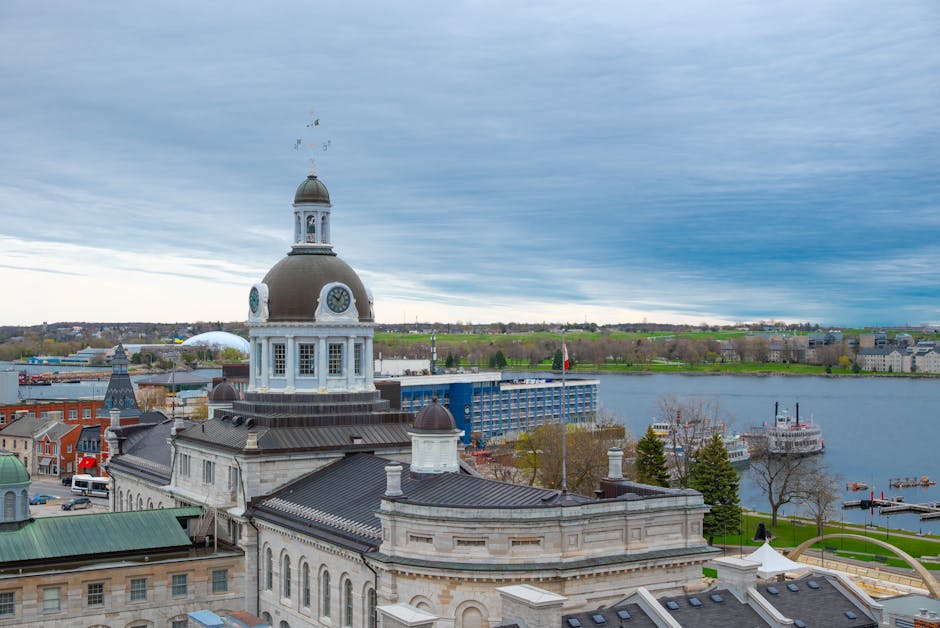 An elevated view of a historic building featuring a large central dome with a clock, topped by a small cupola and weather vane. The building is constructed from light-colored stone with dark roofing materials, and has ornate architectural details along its edges and rooflines. Surrounding structures include other stone buildings with varied roof shapes and colors, some with green patina domes and dark slate roofing. In the background, a wide river stretches across the scene, with a docked white passenger ferry and small boats moored nearby, and a distant shoreline with patches of greenery and scattered buildings. The sky above is filled with blue and gray clouds, suggesting overcast weather, and the environment appears calm, with a mixture of urban and natural elements indicative of a riverside cityscape, relevant to waste management and service accessibility in the area. Rubbish clearance services are implied by the context of waste removal, though no action is depicted in this image.