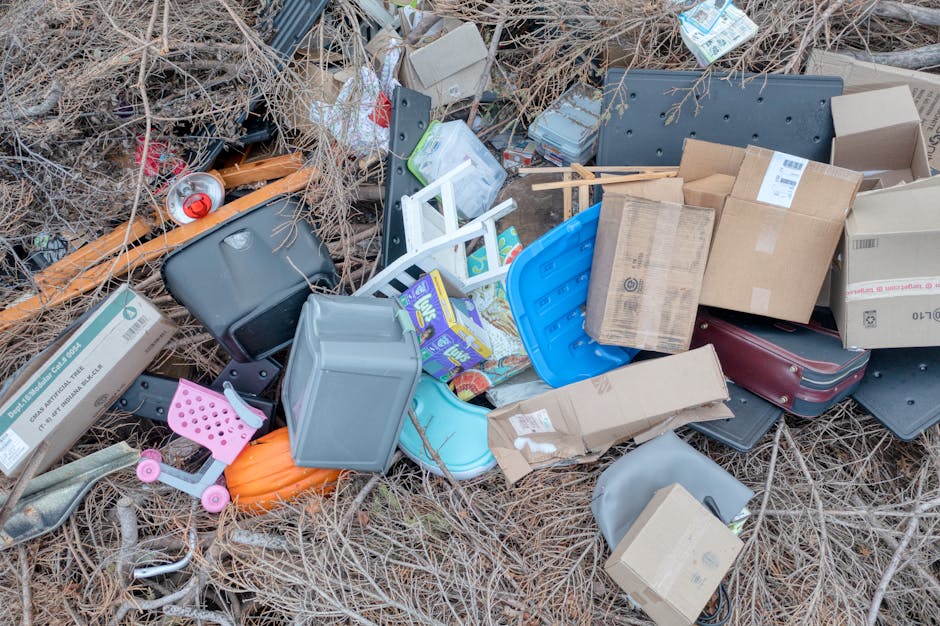 An outdoor scene showing a pile of assorted household rubbish scattered on dry, brown grass and twigs. The collection includes various cardboard boxes, some flattened and others partially expanded, along with plastic containers in different shapes, sizes, and colours such as blue, gray, and pink. A small pink toy scooter is visible among the debris, along with a black plastic case or bag and a partially visible orange item. Several paper and cardboard packages, some with barcodes and labels, are mixed in with the plastics, while a metal can with a red lid lies near the top left. The background consists of dry vegetation, indicating the rubbish is dumped or discarded outside a residential or garden area. Rubbish clearance services like those offered by Rubbish Clearance Kingston could assist with the safe removal and proper disposal of such waste, often involving on-site collection of bulky or multiple waste items, including packaging and household refuse, in accordance with local guidelines and regulations.
