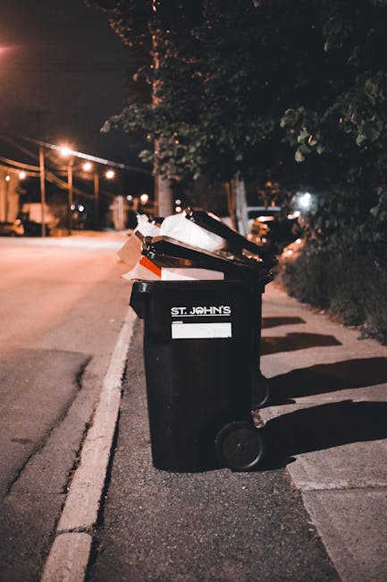 A green old clothing collection box positioned on a sidewalk, surrounded by various discarded items including shoes, socks, clothing, a backpack, and a small suitcase. The box features Chinese characters and symbols indicating it is for donating clothes, shoes, and other small items for reuse. The background consists of foliage and a wooden fence, with additional waste materials scattered in front of the box, such as shoes and small personal belongings. The scene reflects improper disposal of waste, with clutter and rubbish accumulation around the collection point, which is typically used for designated clothing donation purposes but has been overwhelmed by general rubbish. Rubbish clearance services like those from Rubbish Clearance Kingston are often involved in the clean-up and proper disposal of such waste collected from public donation points, ensuring environmental and community cleanliness.