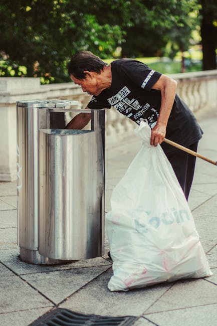 A man with dark hair, wearing a black T-shirt with white text and black trousers, is seen disposing of rubbish into a metal trash bin on a paved outdoor walkway. He is leaning forward with his left arm extended to place waste into the bin, which has a circular opening at the top. The man holds a large white plastic sack filled with rubbish in his right hand, supporting it against his body. The background features a low stone wall and lush green foliage, indicating an outdoor urban park or public space. The lighting suggests a daytime setting with natural light, and the scene reflects routine waste disposal activity, characteristic of independent rubbish collection or on-site clearance, with the man possibly engaged in a community cleanup or personal waste management effort. Rubbish Clearance Kingston performs similar activities, ensuring proper disposal outside of standard council waste services.