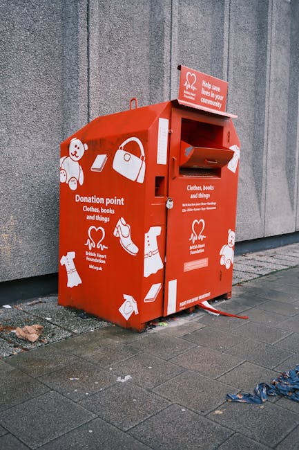 A red donation collection box positioned outdoors on a paved sidewalk against a grey concrete wall. The box is designed for the collection of clothes, books, and other items, with various white graphic icons representing these objects, including a teddy bear, a handbag, a book, and a t-shirt. The front of the box features a slot for donations and a small flap below it, with additional signage indicating that it is a donation point managed by Rubbish Clearance Kingston. The box appears to be made of metal with a textured surface and a glossy finish, and is secured to the ground with red straps. Nearby, the pavement has some small bits of litter, and a discarded blue strap or ribbon can be seen on the right side. The environment suggests a typical urban setting where independent collection points for charitable donations are installed, offering an alternative to regular rubbish removal services and supporting community recycling initiatives.