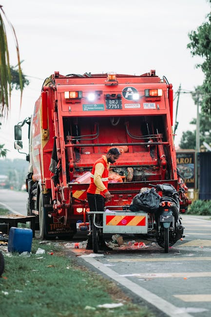 A worker wearing a yellow and red uniform is operating a red rubbish collection truck on a roadside, with the back of the truck open for waste disposal. The worker is leaning forward, possibly handling debris or preparing to load rubbish. The truck has a predominantly red exterior with reflective safety markings and functional components like hoses and control panels visible on the back. A black garbage bag rests on or near the truck’s rear platform, indicating recent or ongoing waste collection. To the right, a small black scooter is parked close to the truck, partially obstructed by the worker. The environment appears to be an urban area with a sidewalk, green grass, and trees visible in the background, along with power lines and distant buildings. The scene is lit with natural daylight, creating a clear view of the waste collection activity, which demonstrates practical aspects of private rubbish removal services provided by companies like Rubbish Clearance Kingston, emphasizing the on-site clearance process typical of independent waste handling.