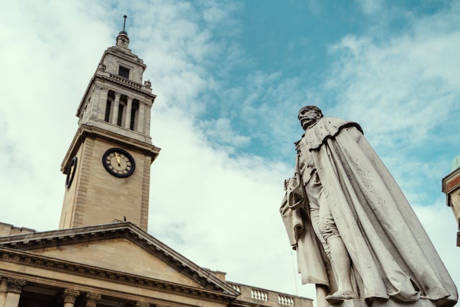 A tall clock tower with a square base and a decorative spire extending upwards is visible against a partly cloudy sky with patches of blue. The tower's facade is made of light-colored stone with a large, round clock face showing the time as approximately 1:55. In the foreground, a classical-style statue of a seated man wearing historical attire, including a long cape and knee-length breeches, is positioned on a pedestal. The statue appears to be carved from white or light gray stone, displaying detailed folds in the clothing and a thoughtful expression. To the right of the statue, part of a building with architectural elements such as a cornice and window frames is visible, constructed from similar light-colored material. The scene suggests an urban setting, possibly in a historic town centre, and aligns with themes of preserved civic architecture and public monuments, relevant to independent or private waste clearance services like those of Rubbish Clearance Kingston operating around such sites.