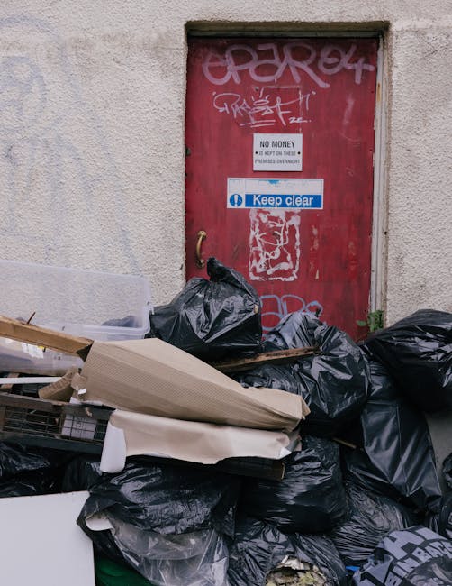 A large pile of black waste bags and discarded packaging materials stacked in front of a red metal door mounted on a beige, textured concrete wall. The waste bags appear to contain household or commercial rubbish and are haphazardly piled, partially covering some cardboard and plastic items. The door, situated within a small framed opening at the top of the wall, features graffiti, a white sign reading 'NO MONEY, NO EXCEPTIONS,' and a blue sticker with white text stating 'Keep clear.' The scene is outdoors, likely on a city or urban street, with natural daylight illuminating the area. This setup suggests a typical roadside rubbish accumulation that might be addressed through private waste collection services, such as those provided by Rubbish Clearance Kingston, to ensure proper removal and disposal outside of council rubbish collection systems.