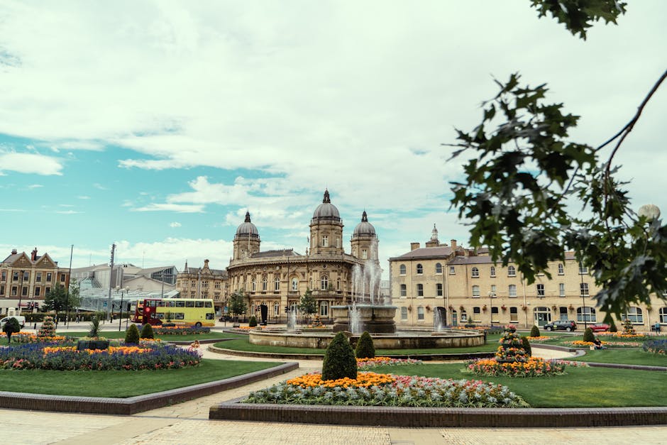 A wide-angle view of a historic city square featuring a large, ornate stone building with dome-shaped towers and detailed architectural elements in the background. In the foreground, there is a circular fountain with multiple water jets shooting upwards, surrounded by well-maintained flower beds filled with colorful, blooming flowers and neatly trimmed small bushes. To the left, a yellow double-decker bus is seen parked on the street, while on the right, neighboring buildings with classic facades line the square. Overhead, the sky is partly cloudy with patches of blue visible, and leafy branches from a tree extend into the top right corner of the image. The scene captures a lively public space with a mix of historic architecture and urban activity, illustrating the environment where private waste management services like Rubbish Clearance Kingston can operate in an organized and professional manner, fitting within a context of alternative rubbish removal options outside of municipal collection.