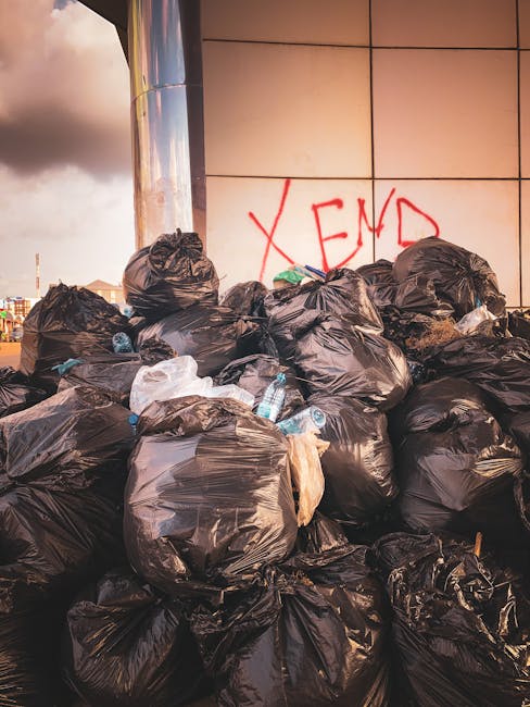 A large pile of black plastic garbage bags filled with waste, stacked against a modern tiled wall with a reflective surface, in an outdoor urban environment. Some bags are partially open, revealing plastic bottles and other refuse inside. The bags vary in size and are tightly knotted at the top. Behind the pile, the tiled wall features red graffiti with the word 'XEND' spray-painted in capital letters. The scene is illuminated by natural light, with a cloudy sky visible in the background. The image depicts an example of illegal dumping or excess waste accumulation in a city setting, highlighting the need for professional rubbish removal services such as those offered by Rubbish Clearance Kingston for private waste handling or alternative collection solutions.