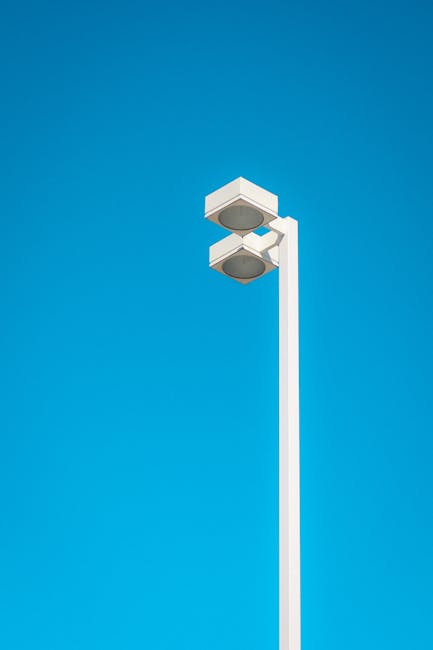 A tall, white street lighting column with two modern, rectangular light fixtures mounted near the top, set against a clear, bright blue sky. The fixtures are enclosed in white casing with dark, circular light sources visible beneath. The pole appears smooth and clean, with no additional attachments or decorations. The scene is outdoor, with no other objects or background elements visible, emphasizing the minimalistic, functional design of the street lamp, which could be seen in urban or suburban areas. This imagery aligns with the idea of well-maintained infrastructure, often associated with environmentally conscious waste management and municipal services, including private rubbish collection and on-site clearance options. The stark contrast between the white pole and the vivid blue sky creates a clear, unobstructed view, highlighting clarity and precision in design, similar to the professional approach of a reliable rubbish removal service like Rubbish Clearance Kingston.