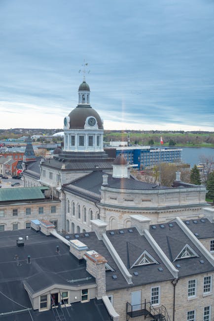 A panoramic aerial view of a historic building with a prominent clock tower featuring a rounded dome and a weather vane on top, situated in an urban area with a mix of older stone structures and modern buildings. The foreground includes rooftops with dark shingles, chimneys, and small dormer windows, some of which have communication cables attached. Behind the building, there are trees and open green spaces leading to a river or water body, with a partly cloudy sky overhead. The scene captures the contrast between traditional architecture and surrounding development, reflecting a setting where independent disposal and private waste handling by services like Rubbish Clearance Kingston may be relevant to maintaining the area’s appearance and managing rubbish collected from historic and modern structures.