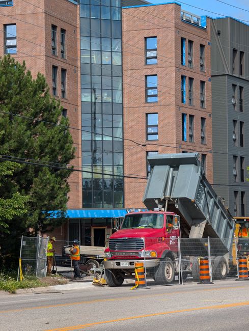 A red industrial dump truck is positioned in front of a modern multi-storey building with a brown brick façade and large glass windows. The truck's open-top container is raised at an angle, suggesting loading or unloading activity, and the vehicle appears to be engaged in site work or waste removal tasks. Surrounding the truck is a safety barrier consisting of metal fencing and orange and black striped bollards, indicating an active work zone. Several workers wearing high-visibility vests and helmets are present near the building entrance, possibly coordinating or overseeing the operation. The environment includes a sidewalk, some greenery with a large tree on the left, and overhead electrical wires crossing the scene. The lighting suggests daytime with natural, diffuse sunlight, and the scene reflects a typical construction or building maintenance context, potentially related to on-site waste removal or private disposal operations managed by Rubbish Clearance Kingston, aligning with alternative rubbish collection services.