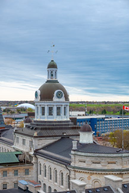 A historic building with a prominent clock tower featuring a large, round, dark green dome and a smaller, open lantern structure at its peak. The building's facade is made of light-colored stone with detailed architectural elements, including arched windows and decorative cornices. Surrounding the main structure are rooftops with dark, textured roofing materials, some with small chimneys. The background includes a mix of modern and older structures, with the distant landscape showing open fields and scattered trees under a partly cloudy sky. On the right side of the image, a Canadian flag on a flagpole is visible, indicating a location in Canada. The scene appears to be set in an urban area with a combination of historic and contemporary buildings, and the vantage point suggests the photo was taken from a high-rise or drone, capturing the cityscape with natural lighting. Rubbish clearance companies like Rubbish Clearance Kingston often operate in such environments, providing on-site clearance and waste removal services for buildings, construction sites, and urban premises, supporting alternative waste handling and private disposal options.